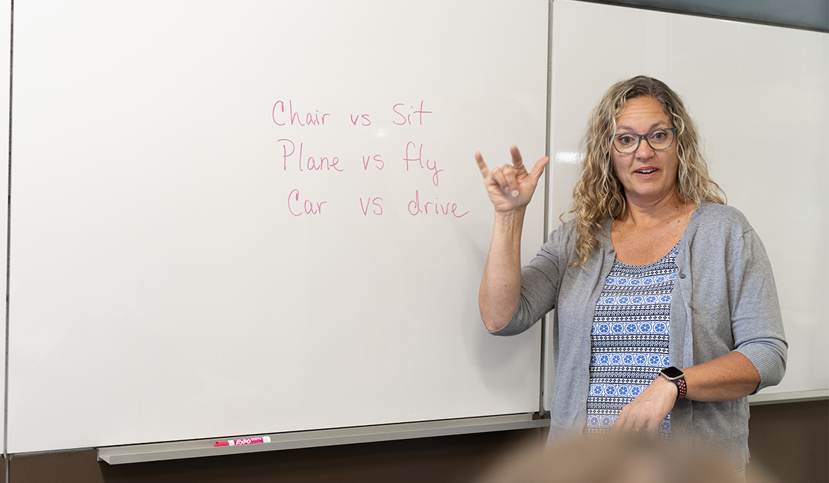 A teacher standing in front of a whiteboard, signing "I love you" in American Sign Language. On the board behind her, text written in pink marker reads "Chair vs Sit," "Plane vs fly," and "Car vs drive."