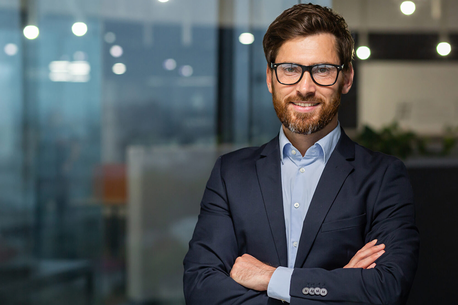 Smiling man in glasses and a navy suit stands with arms crossed in an office