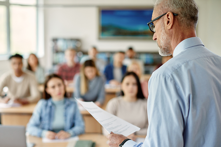 A professor holding papers addresses a classroom full of attentive students.