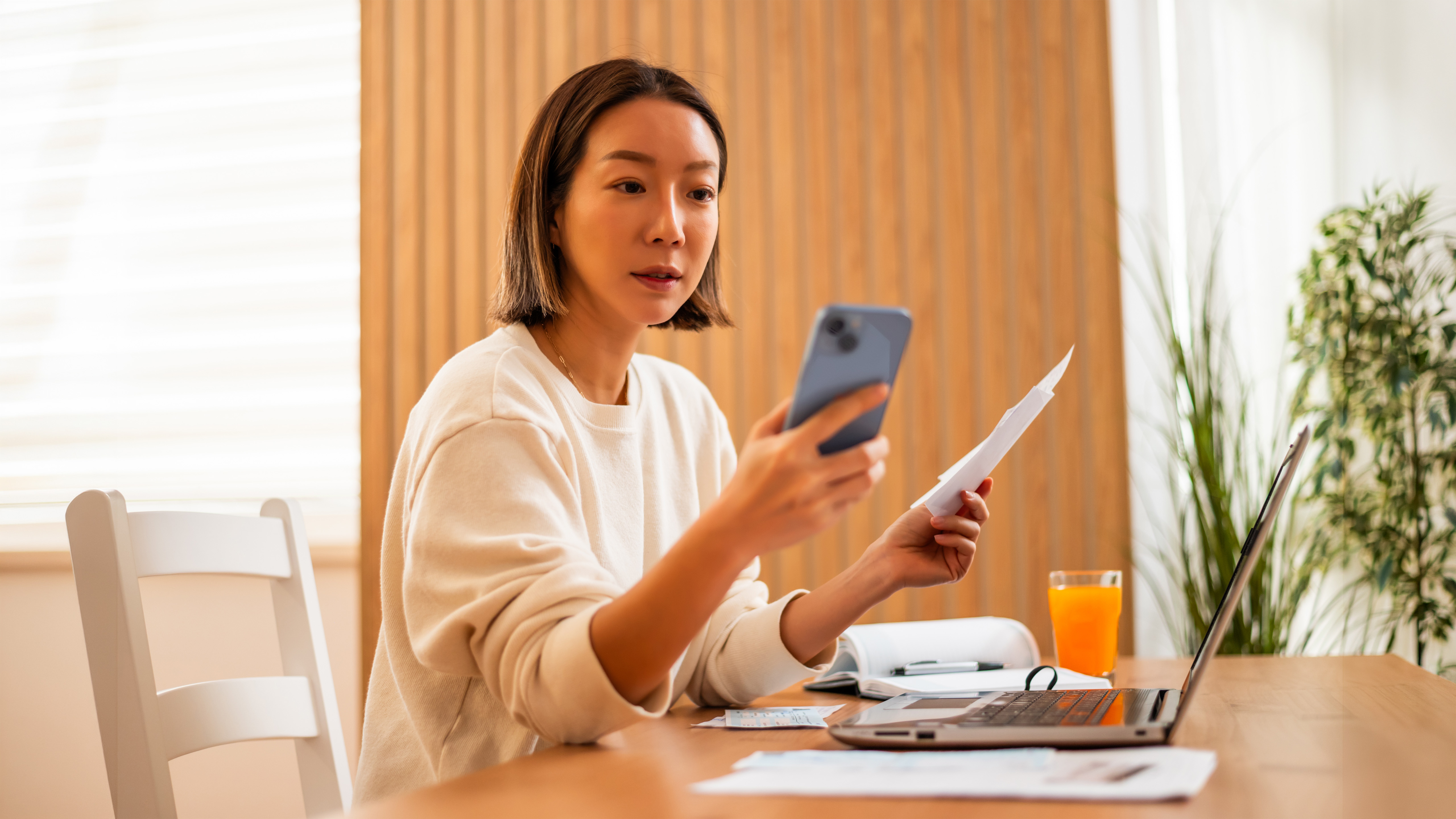 A woman sits at a table with a laptop and paperwork, holding a smartphone and a document as she reviews her finances at home.