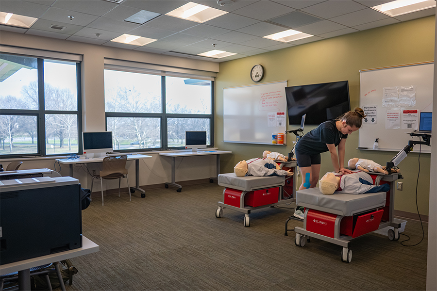 A brightly lit makerspace and simulation lab. In the foreground, a student in a black shirt practices CPR on a medical mannequin resting on a specialized mobile cart. The room is equipped with dual-monitor computer stations, whiteboards, and large windows overlooking a campus landscape.