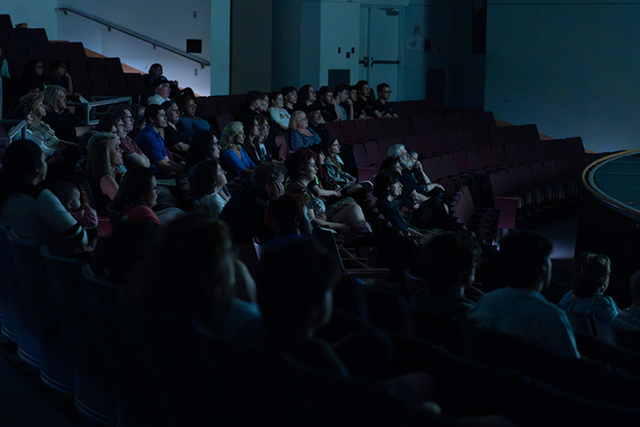 A large audience sits in the tiered seating of the JBK Theater, watching a presentation or film in the dimmed auditorium.