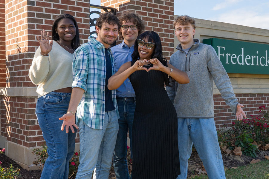 Five smiling students pose together outdoors in front of a brick entrance sign that reads "Frederick."