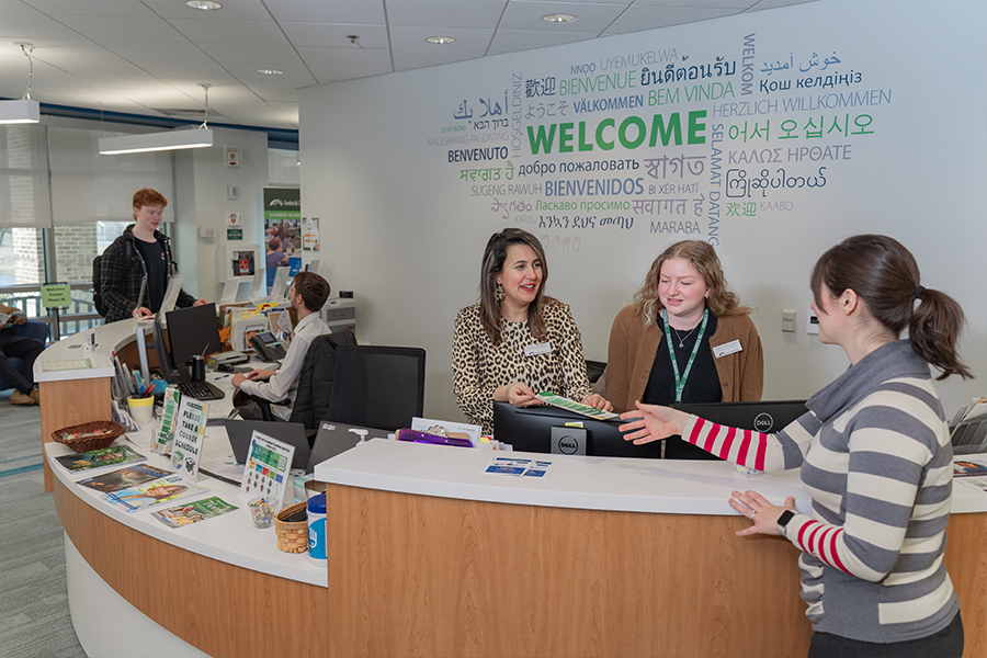 Staff members at a curved white welcome desk assist a student in a brightly lit office. Behind them, a large wall graphic features the word WELCOME in many different languages. The scene is professional, helpful, and inclusive.