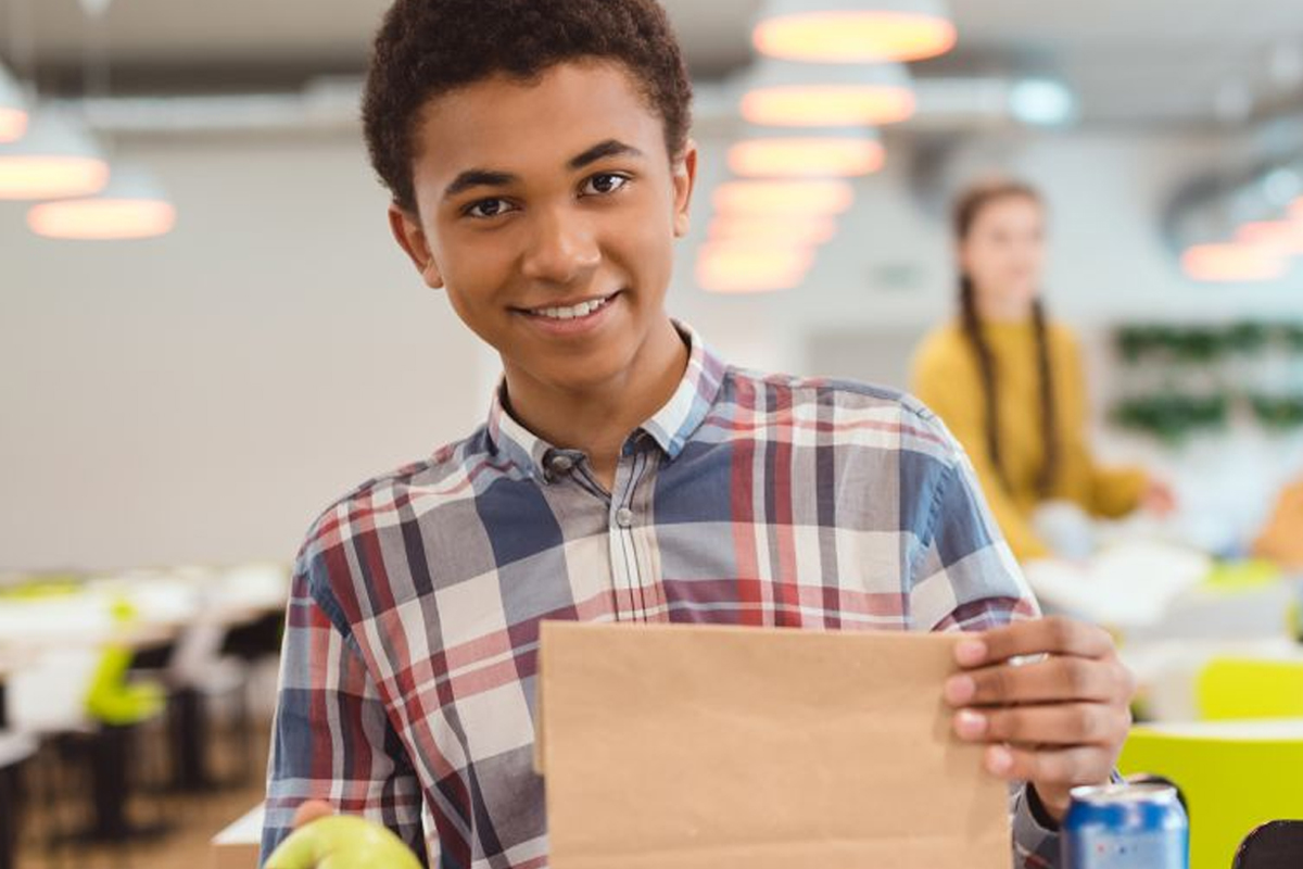 A smiling teenage boy holds a brown paper lunch bag in a school cafeteria, with an apple and a drink can on the table in front of him and other students visible in the background.