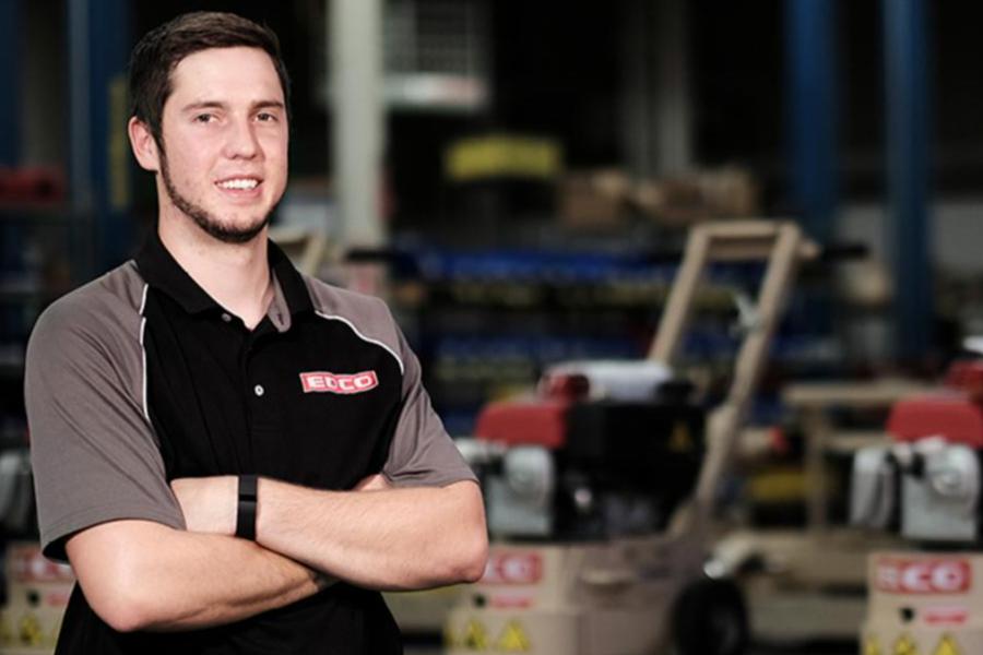 A young man in a branded polo shirt standing with arms crossed in an industrial warehouse setting.
