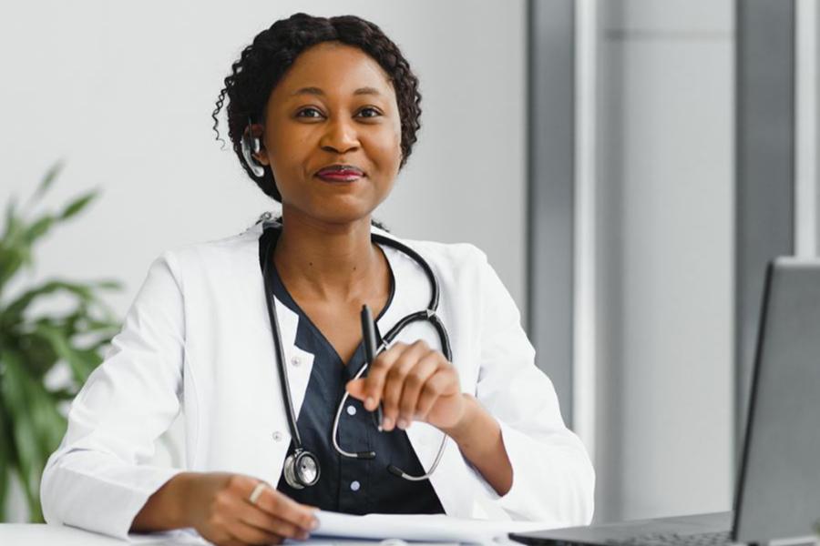 A professional portrait of a female doctor in a white lab coat with a stethoscope around her neck, wearing a hands-free headset while working at a laptop.