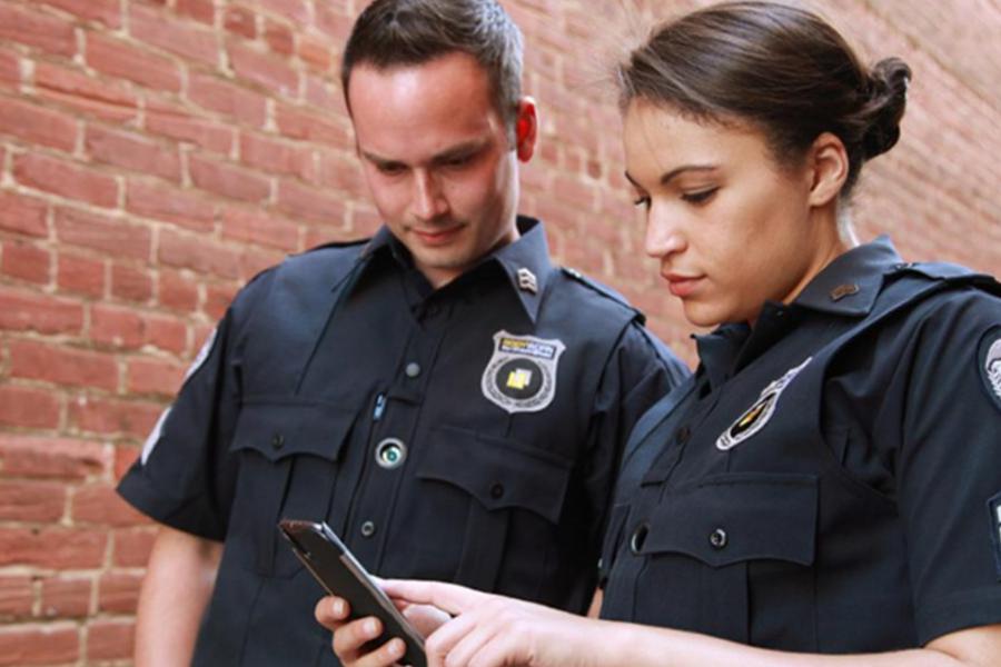 Two law enforcement officers in dark blue uniforms stand against a brick wall while looking at a smartphone together.