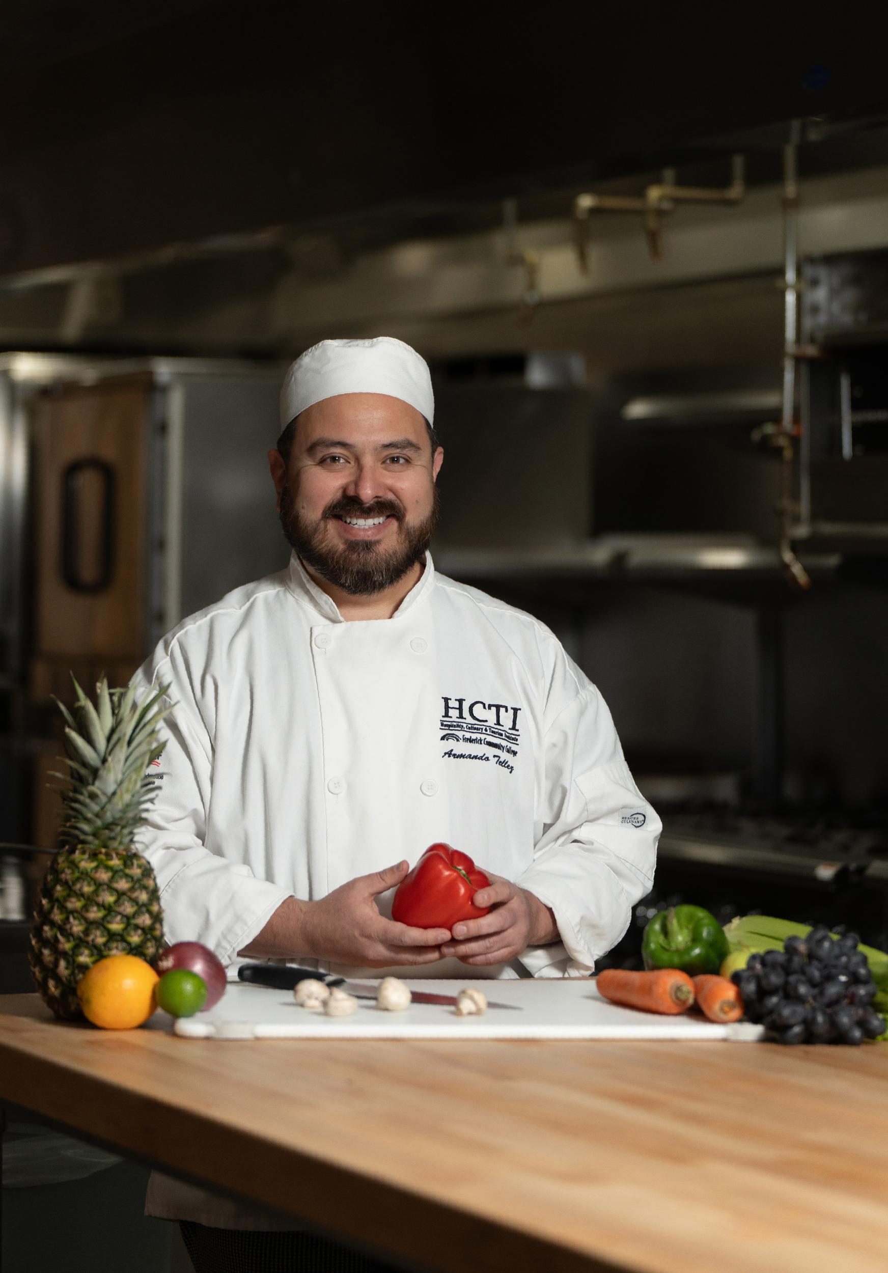 man in white chef coat with HCTI logo on chest, holding a red pepper. A pineapple and vegetables are on the counter.